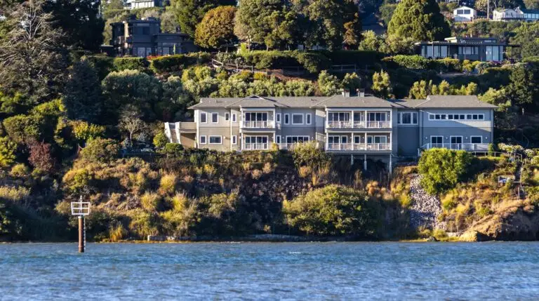 A scenic view of a large waterfront mansion surrounded by trees and greenery, overlooking the sea.